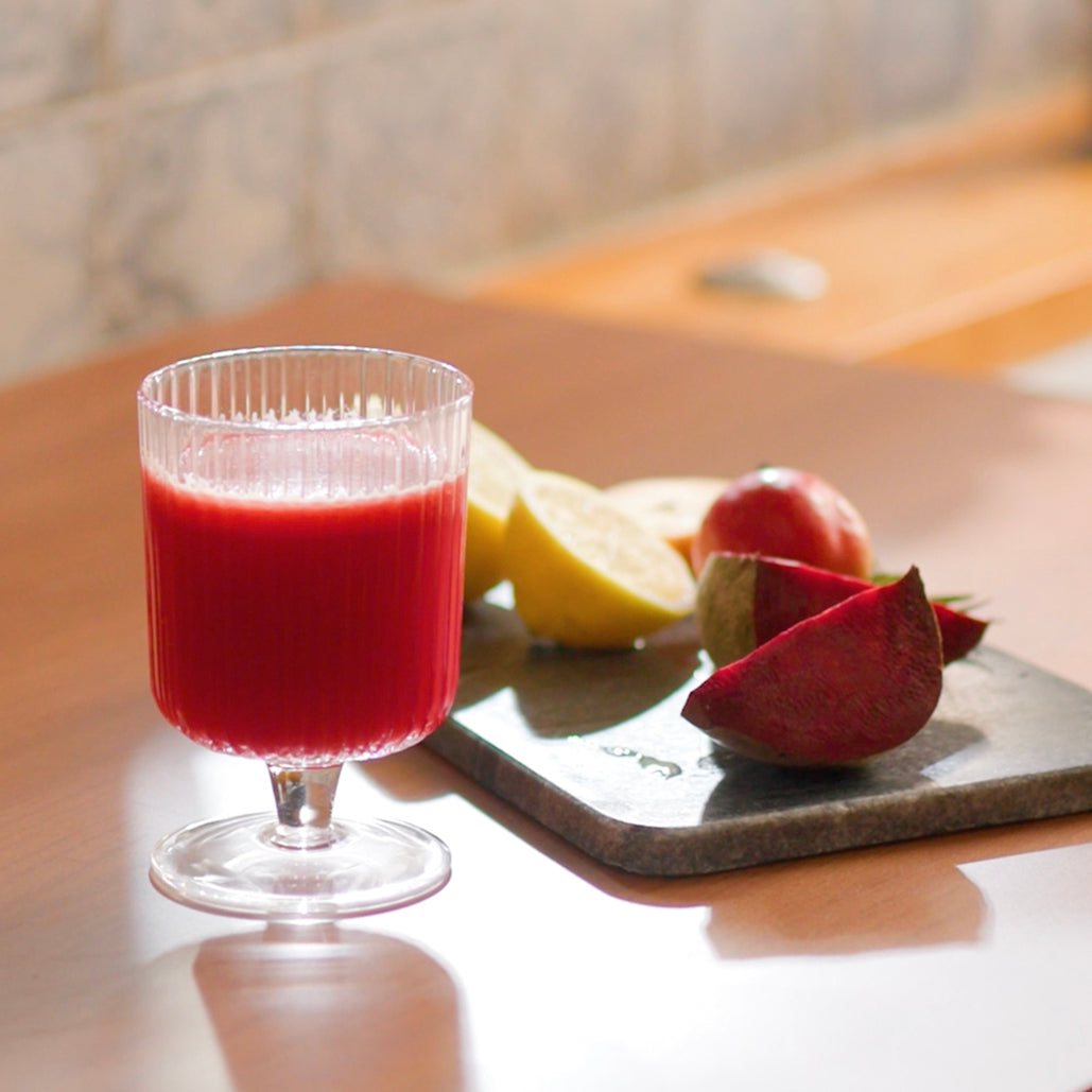 A glass of bright red juice on a table with sliced beet, lemon, and an apple placed beside it.