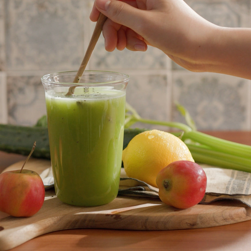 A glass of green juice being stirred with a spoon, with apple, lemon, cucumber, and celery placed around it.