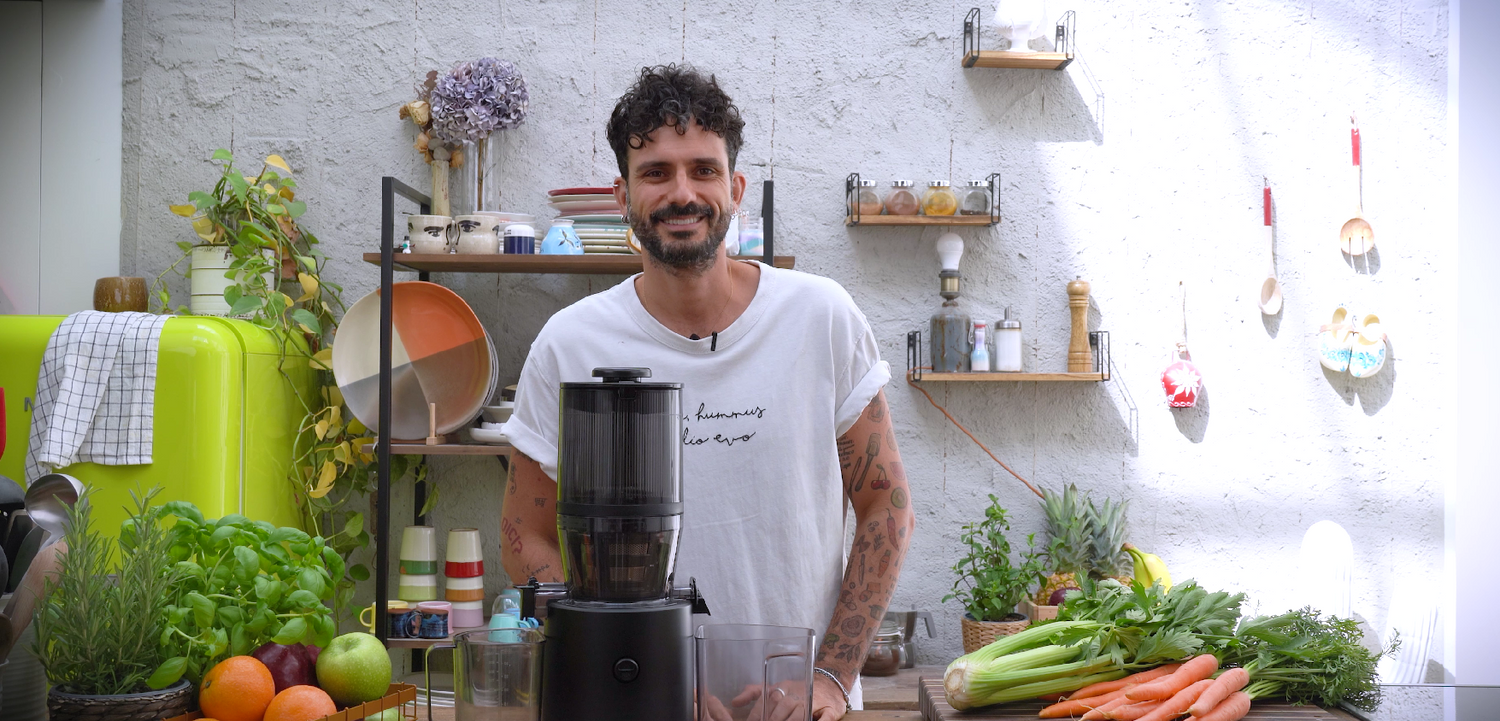 Person standing behind a juicer on a kitchen counter with fruits and vegetables