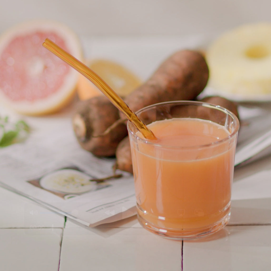 Glass of light orange juice with a straw, with grapefruit and root vegetables in the background