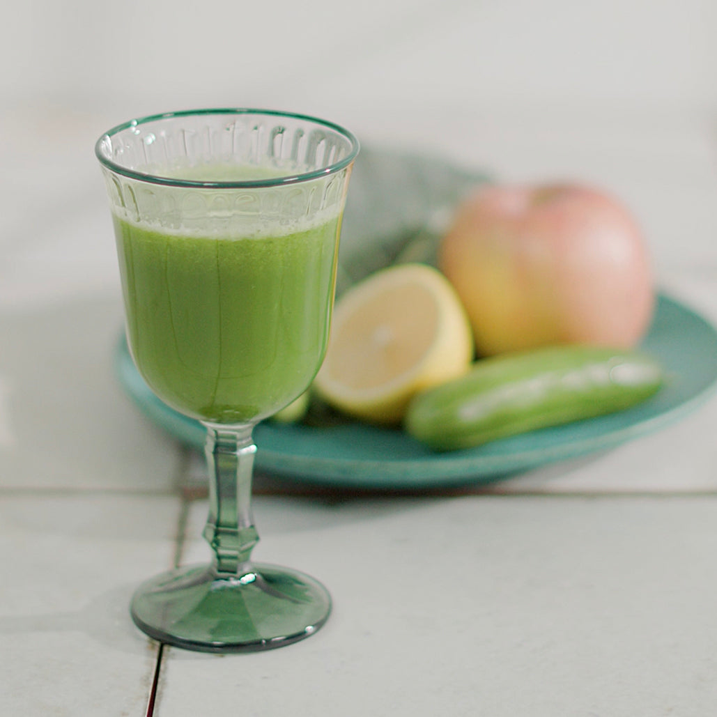 Glass of green juice with an apple, lemon, and cucumber on a plate in the background