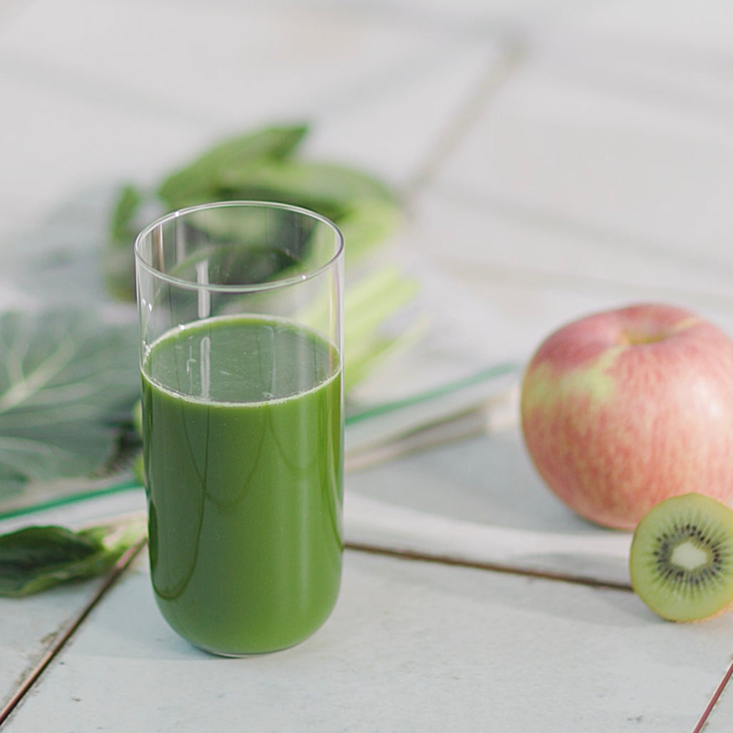 Glass of green juice with an apple, kiwi, and leafy greens on a table