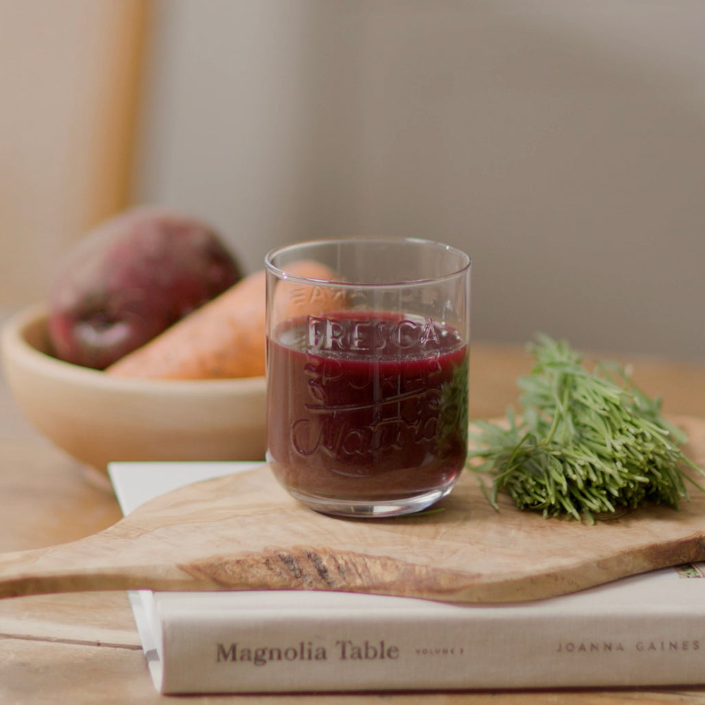 Glass of dark red juice with carrot, beet, and wheatgrass on a wooden board