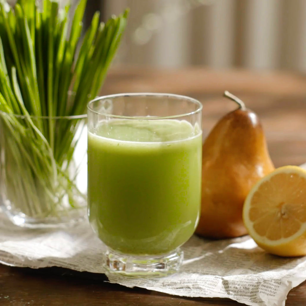 A glass of green juice on a table, with fresh wheat grass, a pear, and a sliced lemon beside it.