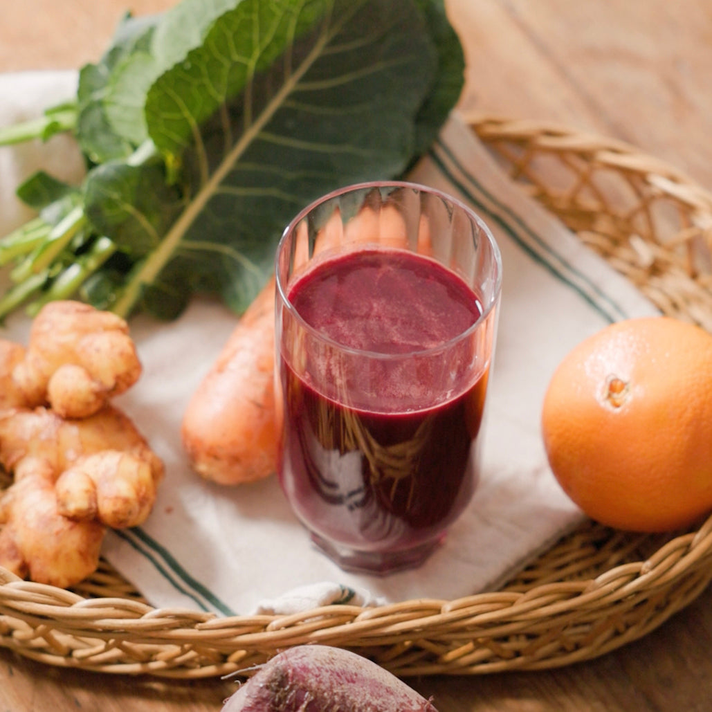 A glass of deep red juice placed on a tray with fresh ingredients including ginger, kale, carrot, orange, and beet.