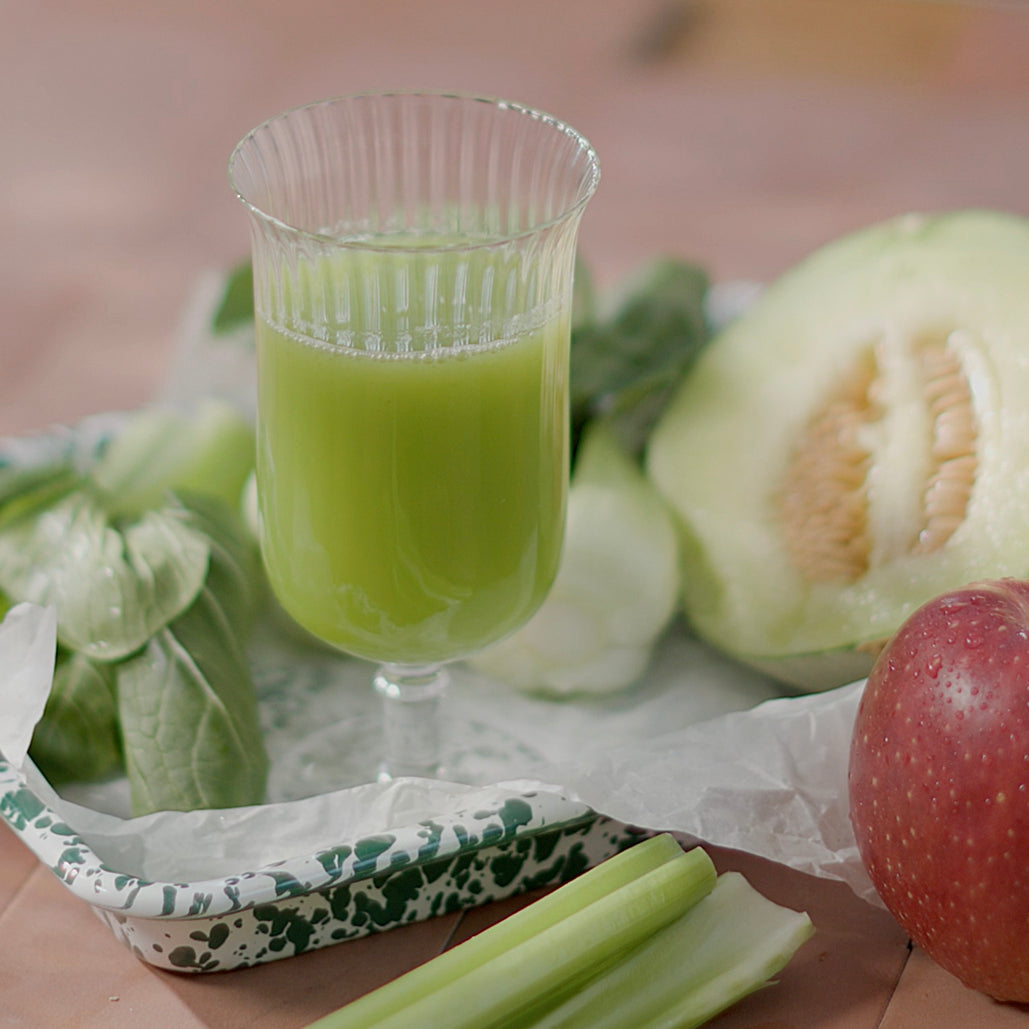 A glass of light green juice on a tray with fresh melon, apple, bok choy, and celery arranged around it.