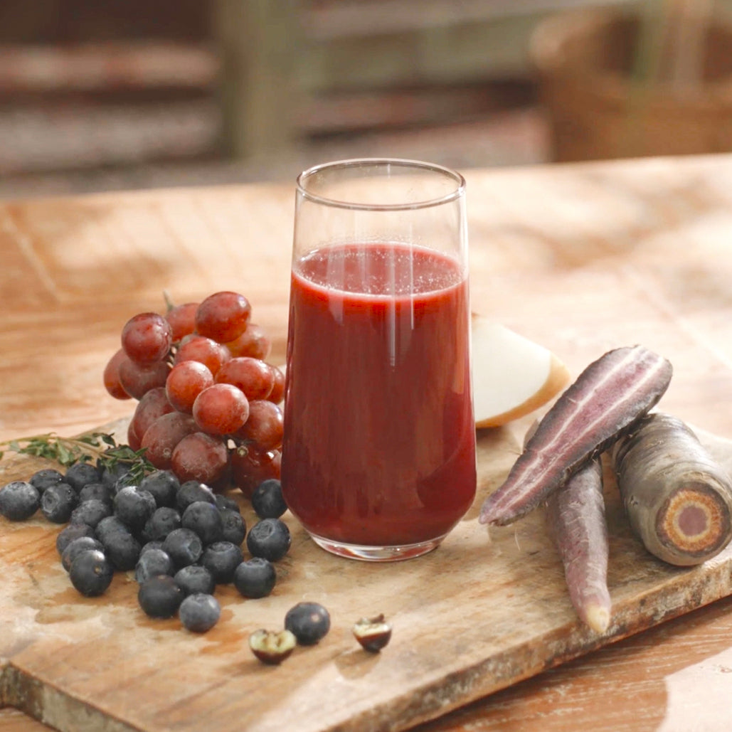 Glass of dark red juice with grapes, blueberries, and carrots on a wooden board