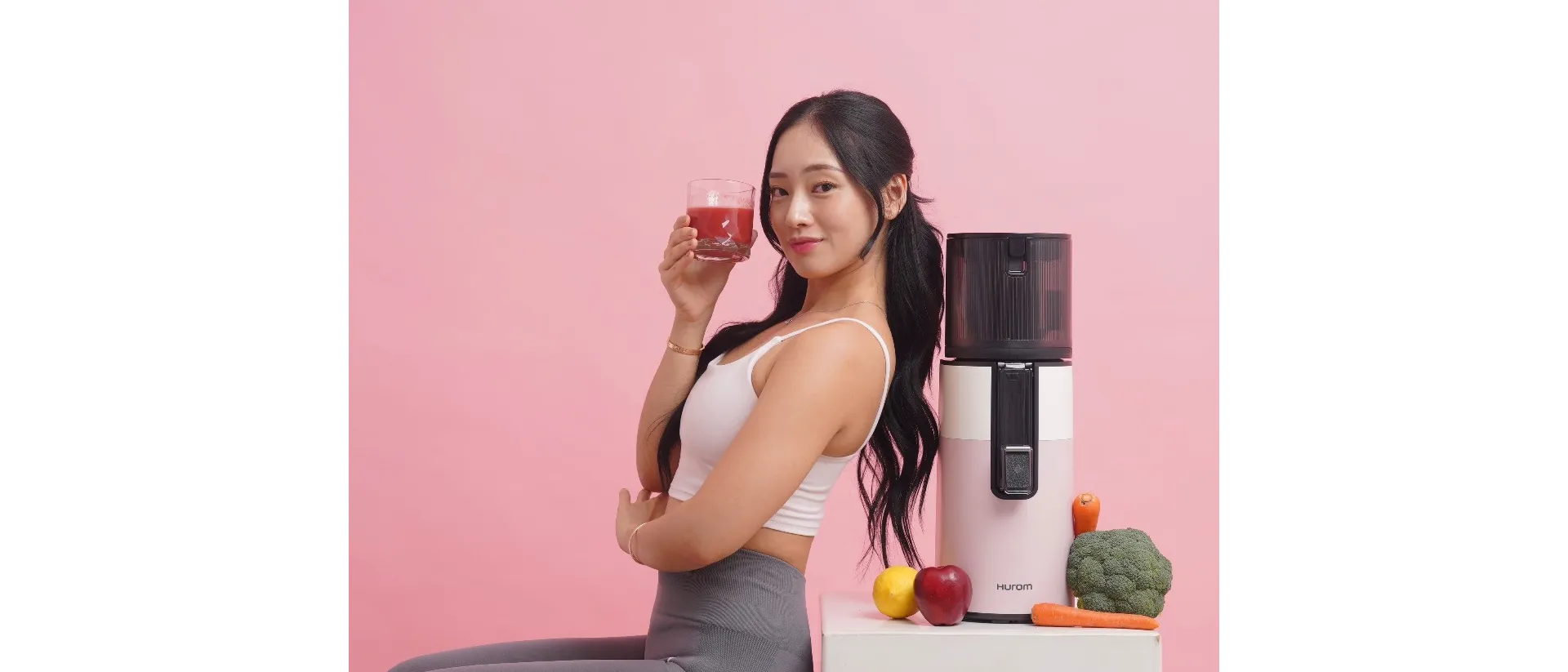 Person holding a glass of juice next to a juicer with fruits and vegetables on a table