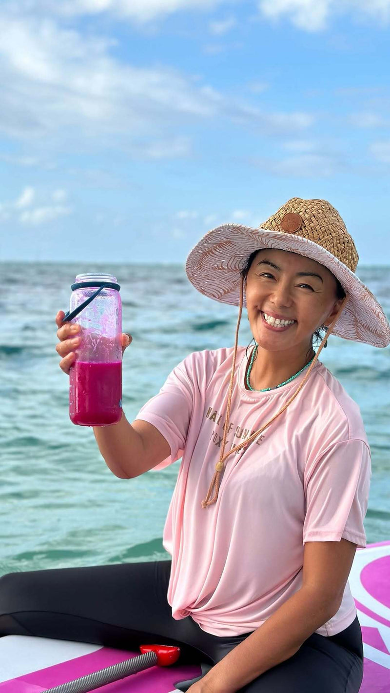 Person holding a pink drink on a paddleboard with ocean and sky in the background