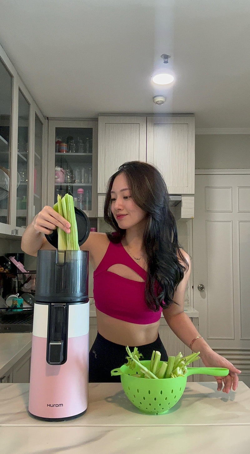 Woman in a kitchen holding a pink and black juicer and a green colander with celery sticks.