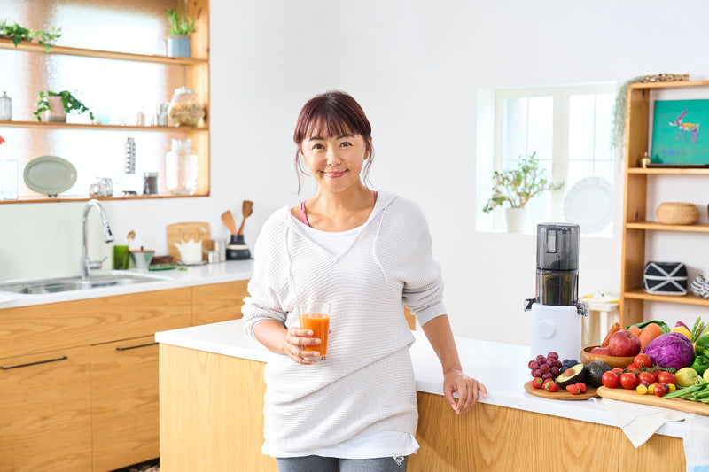 Woman holding a glass of juice in a kitchen with fruits and vegetables on the counter.