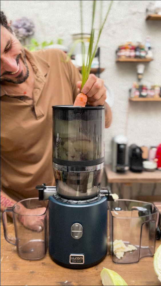 Person using a juicer with a carrot and green leafy vegetable in a kitchen setting.