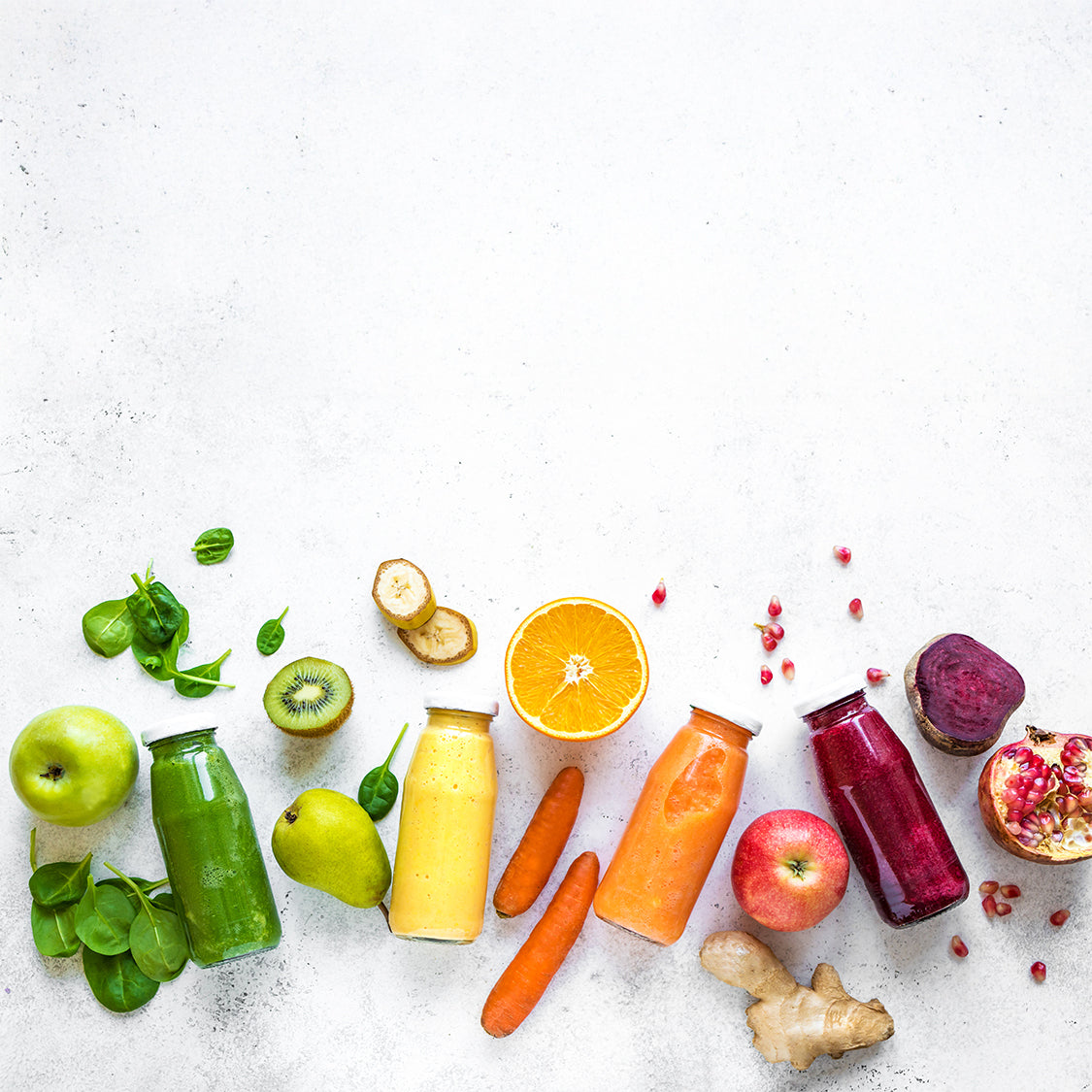 Assorted fruits, vegetables, and bottles of green, yellow, orange, and red juice on a light gray surface.