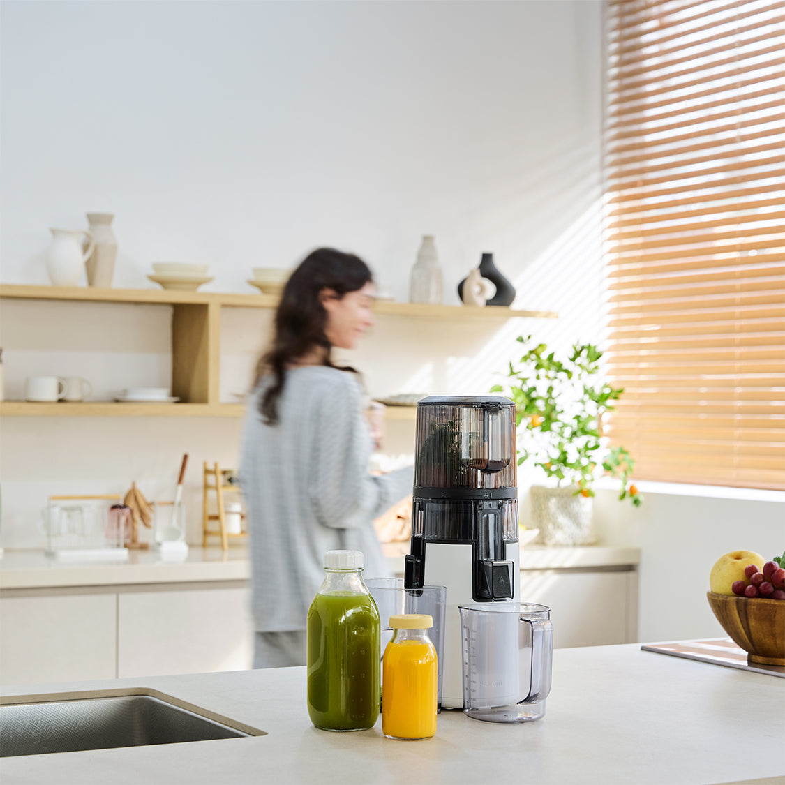 Kitchen scene with a person in the background, a juicer on the counter, and bottles of juice.