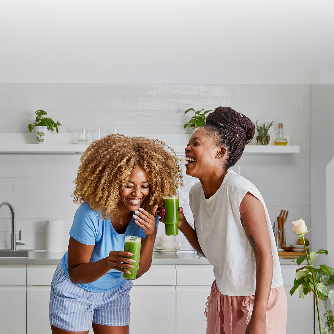 Two women in a kitchen holding green juices and laughing.