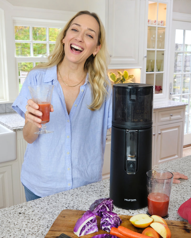 Woman in a kitchen holding a glass of juice next to a black juicer.
