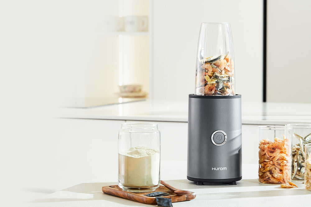 Black Hurom blender on a kitchen counter with ingredients inside, next to a glass of liquid. Power Blender BL-D01