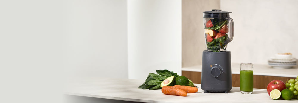 Blender with ingredients on a kitchen counter, next to a glass of green juice. BL-E01
