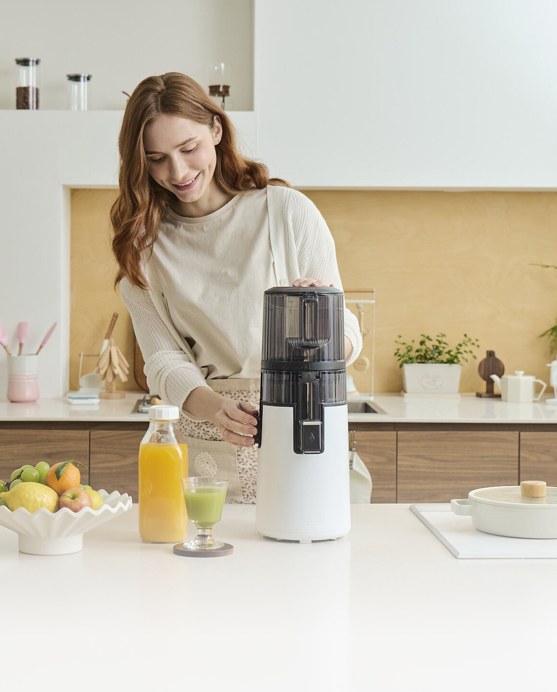 A smiling woman in a bright, modern kitchen using a white Hurom slow juicer to prepare fresh juice. On the counter, there are bottles of orange and green juice alongside fresh fruits, illustrating Hurom's story of making healthy living easy and enjoyable through "Care & Use."