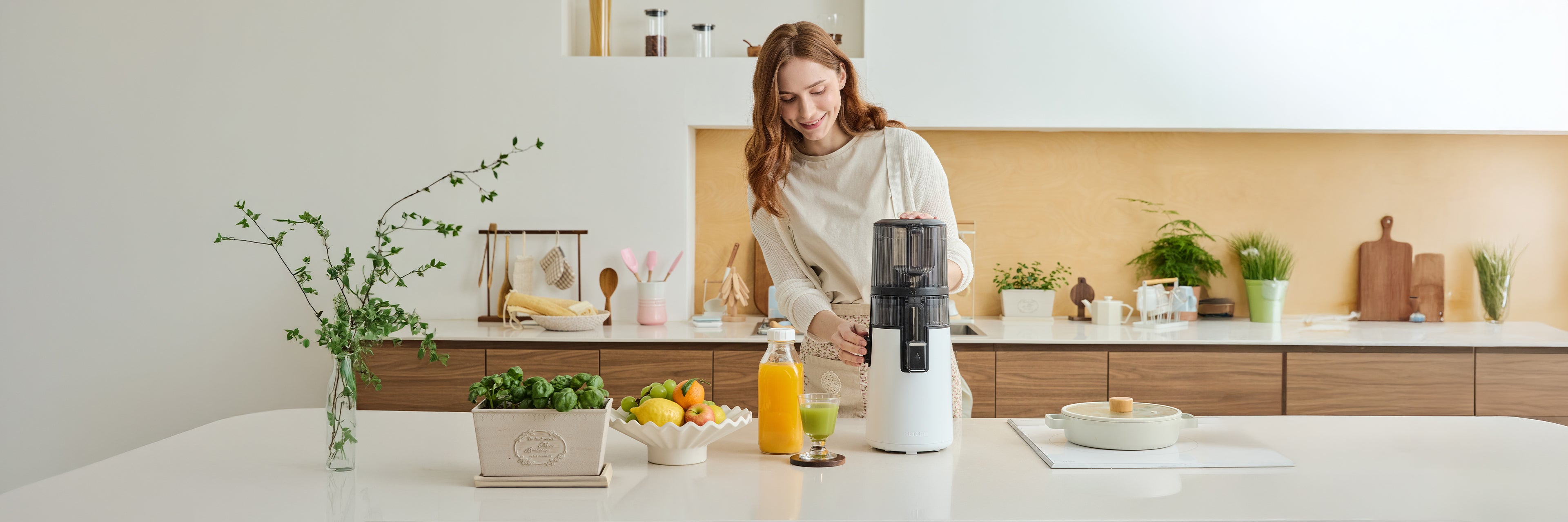 A smiling woman in a bright, modern kitchen using a white Hurom slow juicer to prepare fresh juice. On the counter, there are bottles of orange and green juice alongside fresh fruits, illustrating Hurom's story of making healthy living easy and enjoyable through "Care & Use."