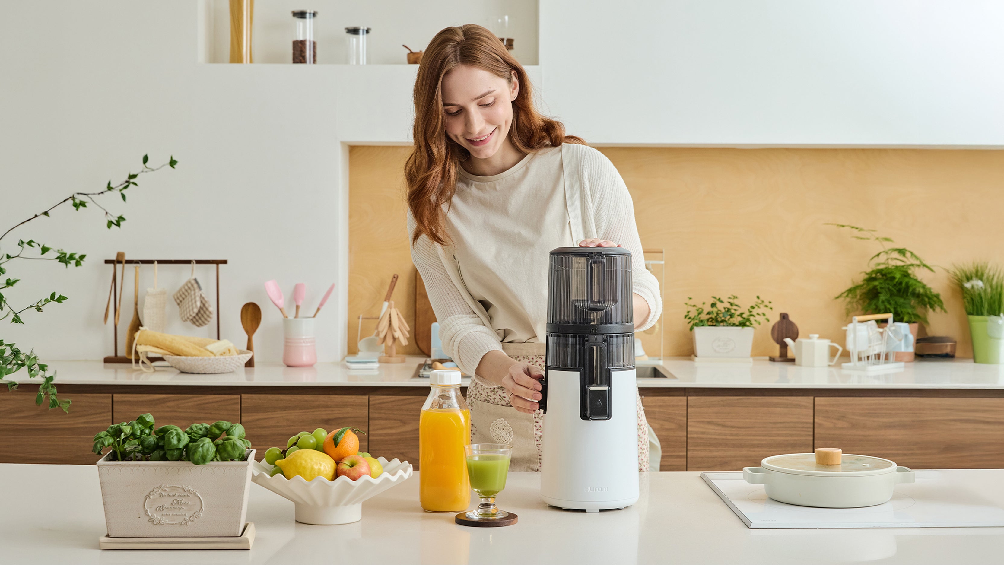A smiling woman in a bright, modern kitchen using a white Hurom slow juicer to prepare fresh juice. On the counter, there are bottles of orange and green juice alongside fresh fruits, illustrating Hurom's story of making healthy living easy and enjoyable through "Care & Use."