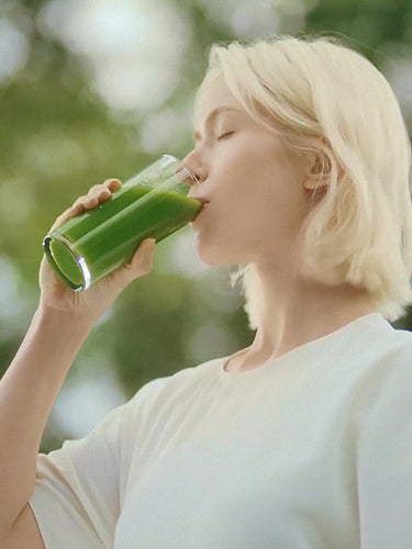 A young woman with her eyes gently closed is drinking vibrant green juice from a glass outdoors, bathed in warm sunlight.